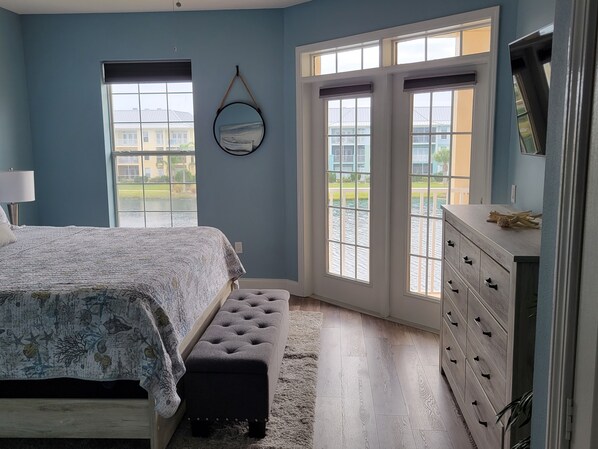 A bedroom with a bed, a bench, a dresser, and a large window with a view of buildings and water.