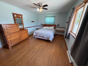 Une chambre à coucher avec une commode en bois, un lit recouvert d’une courtepointe blanche, un ventilateur de plafond et une fenêtre avec des rideaux.