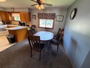 Une cuisine avec des meubles en bois, une table à manger avec des chaises, un ventilateur de plafond et une fenêtre donnant sur un beau paysage.