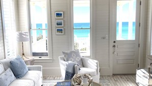 Una habitación luminosa y blanca con vistas al océano, que incluye un sofá blanco, una silla de mimbre y una mesa de centro redonda con objetos decorativos.