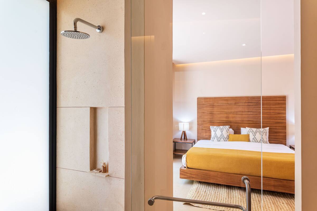A modern hotel room with a large bed, a wooden headboard, a nightstand, and a shower with a handheld showerhead.