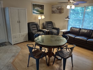 A living room with a glass-top table surrounded by four chairs, a brown leather sofa, and a white cabinet.