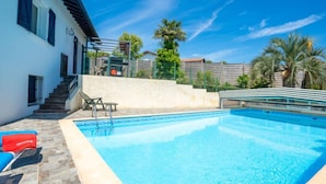 A swimming pool with a tiled deck, a staircase leading to a covered area, and a white building with dark shutters.