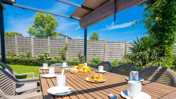 An outdoor wooden table with chairs, a fruit basket, and plates with cups, set in a sunny patio area with a wooden fence and greenery in the background.