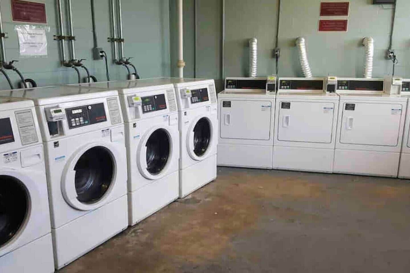 A row of white front-loading washing machines in a commercial laundry room with visible pipes and ventilation ducts on the walls.