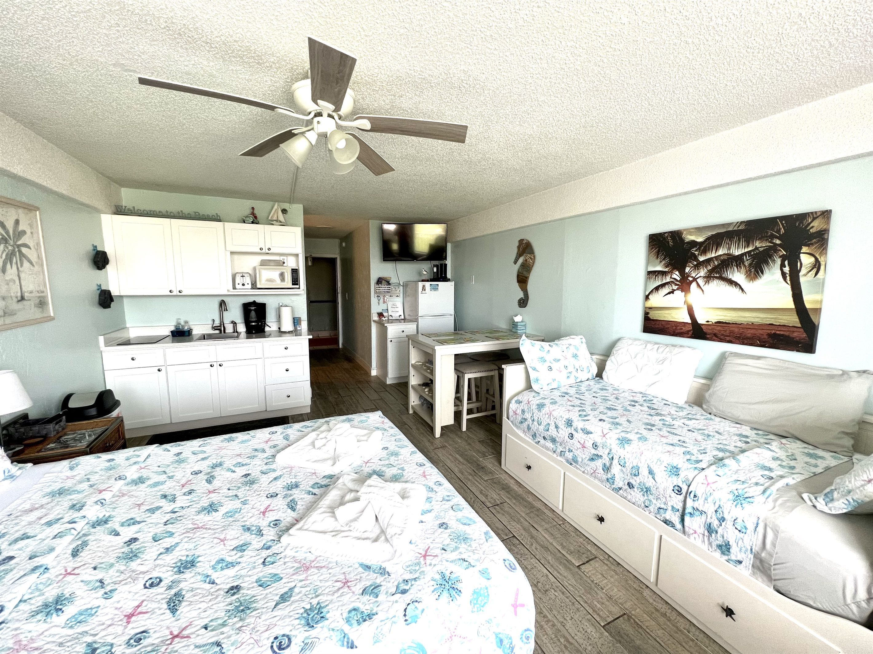 A room with two beds, a kitchenette, a ceiling fan, and a painting of a beach scene.