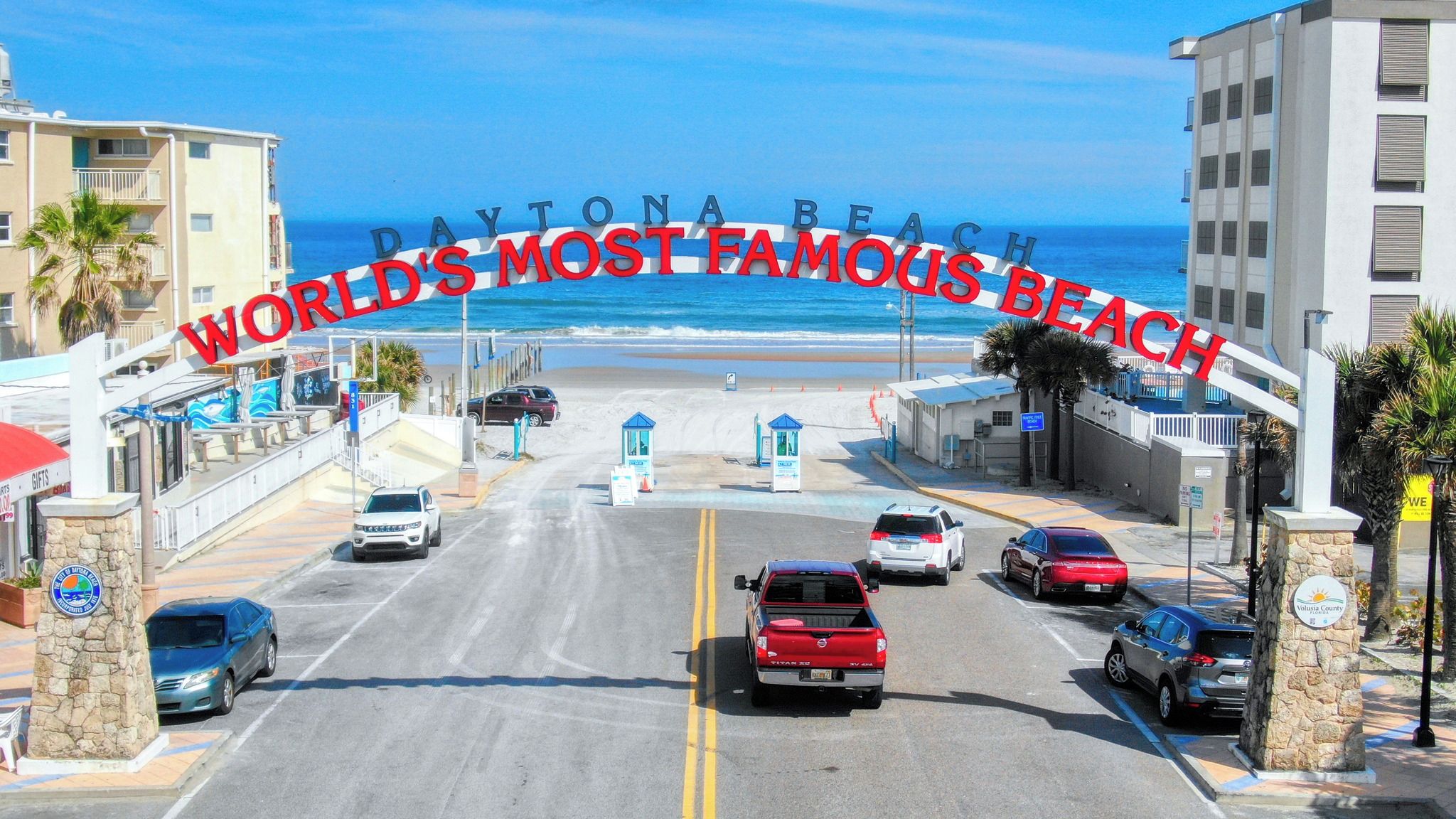 A street view of Daytona Beach with a clear view of the ocean, beach, and a parking area with cars.