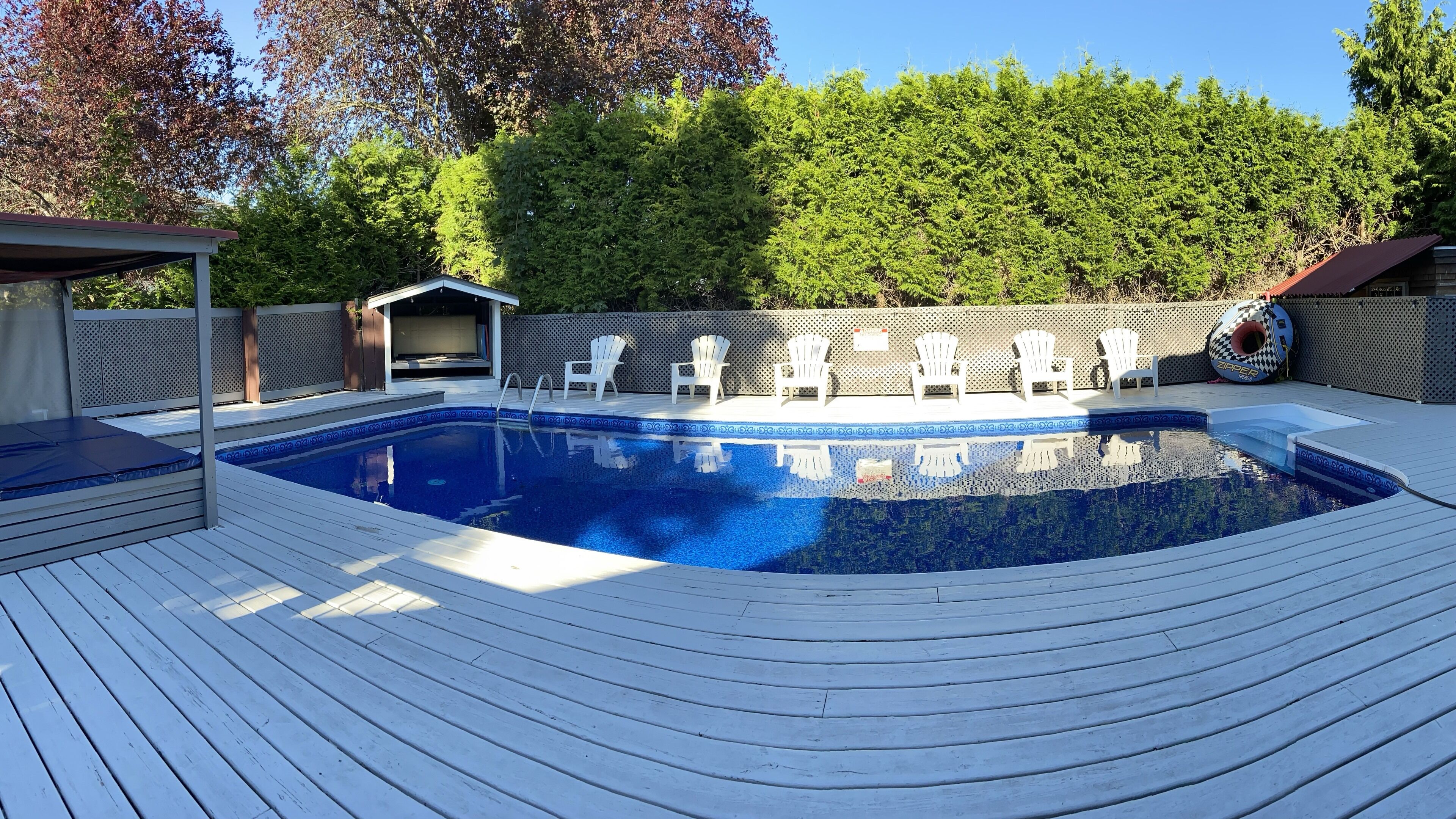 A rectangular swimming pool with a wooden deck, surrounded by white Adirondack chairs, a covered seating area, and a fence with trees in the background.