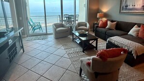 Uma sala de estar espaçosa com uma grande porta de vidro deslizante que dá para uma varanda com vista para o oceano. Há um sofá, uma mesa de centro e um espaço de jantar com cadeiras.
