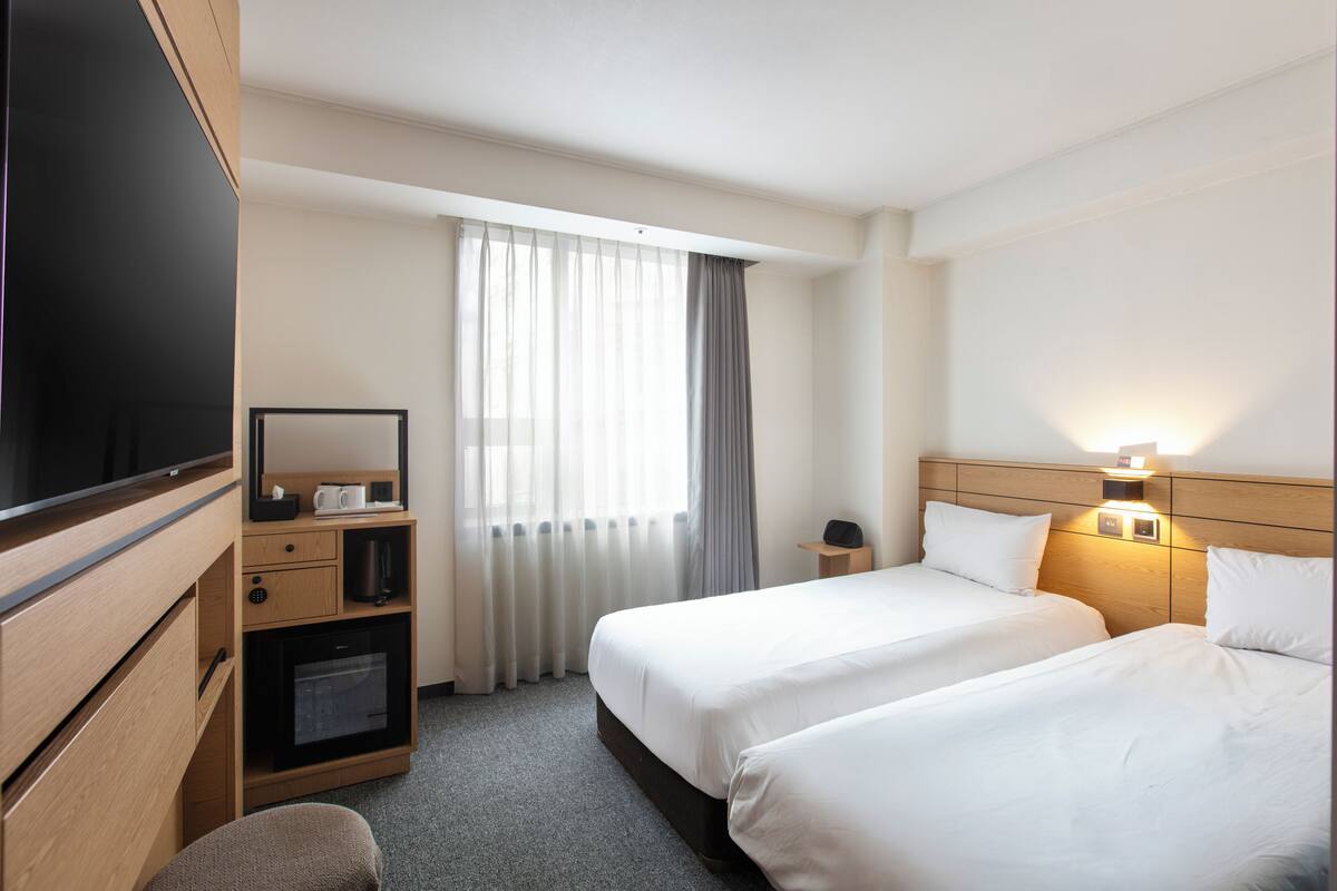A hotel room with two beds, a TV, a desk with a coffee maker, and a window with curtains.