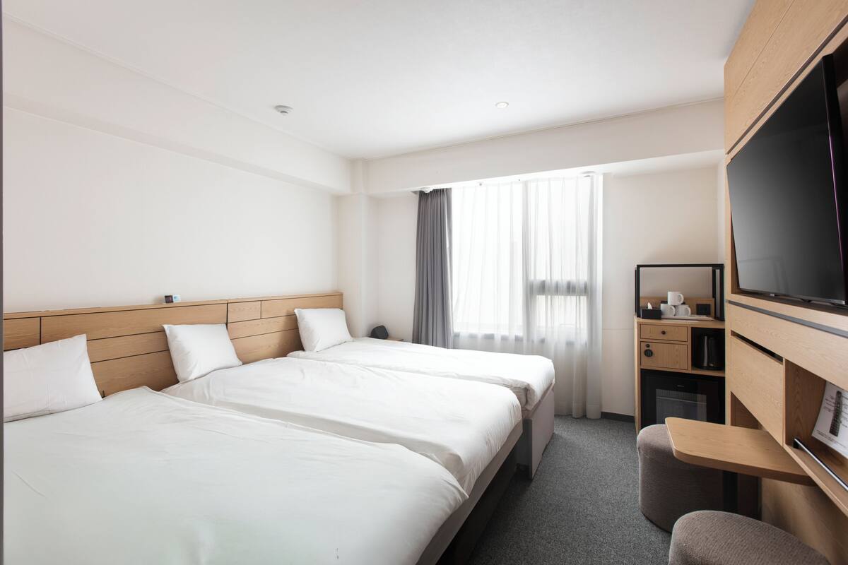 A hotel room with two beds, a desk, a TV, and a window with curtains.