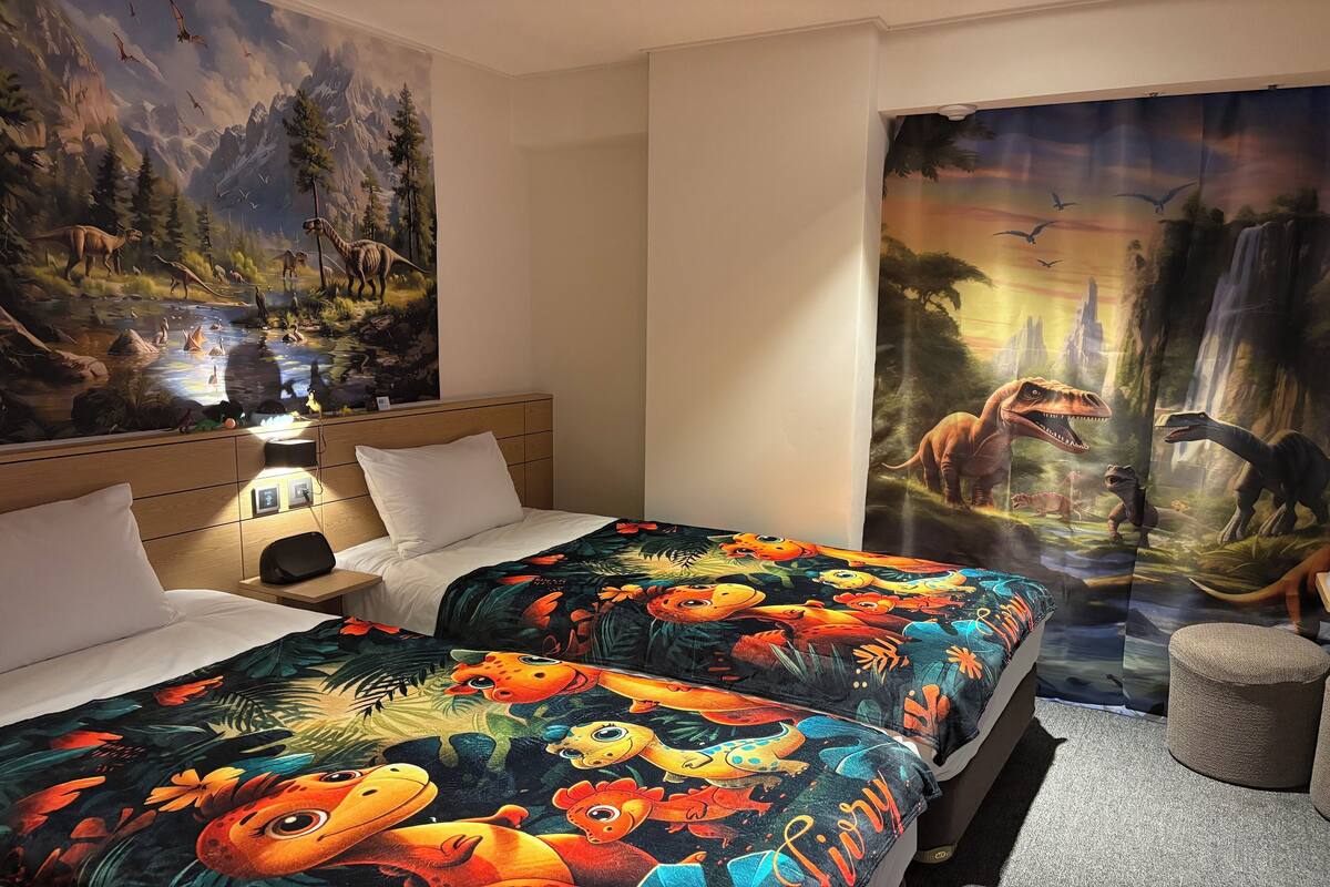 A hotel room with two beds, a mural of dinosaurs on the wall, and a large window with a view of a waterfall.