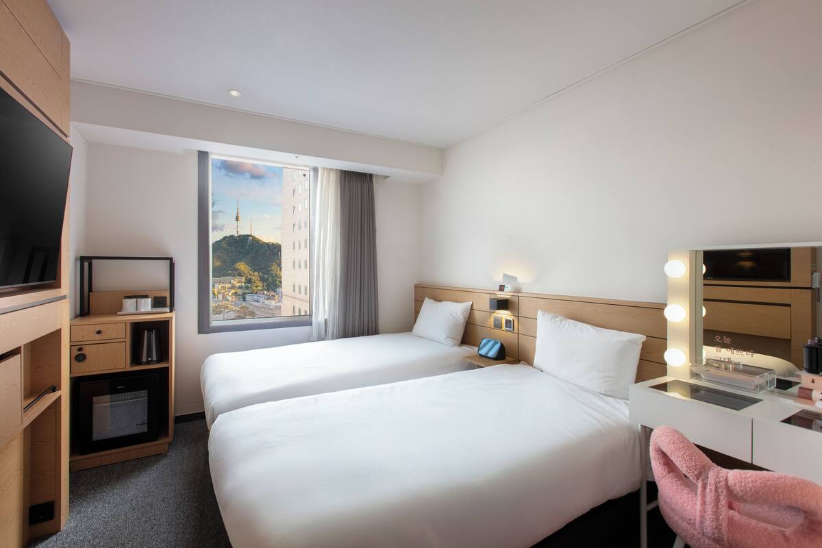 A hotel room with two beds, a TV, a desk with a mirror, and a view of a cityscape.