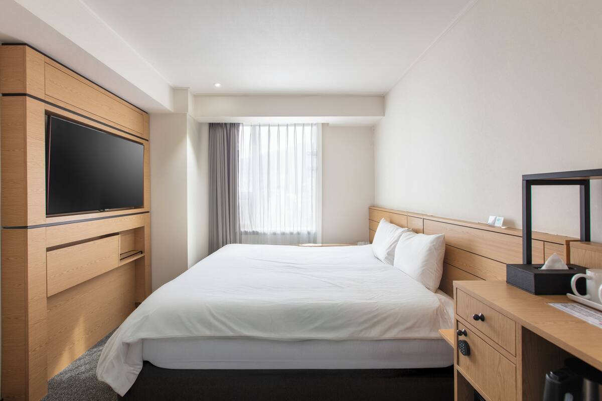 A hotel room with a bed, a TV, a desk, and a window with curtains.