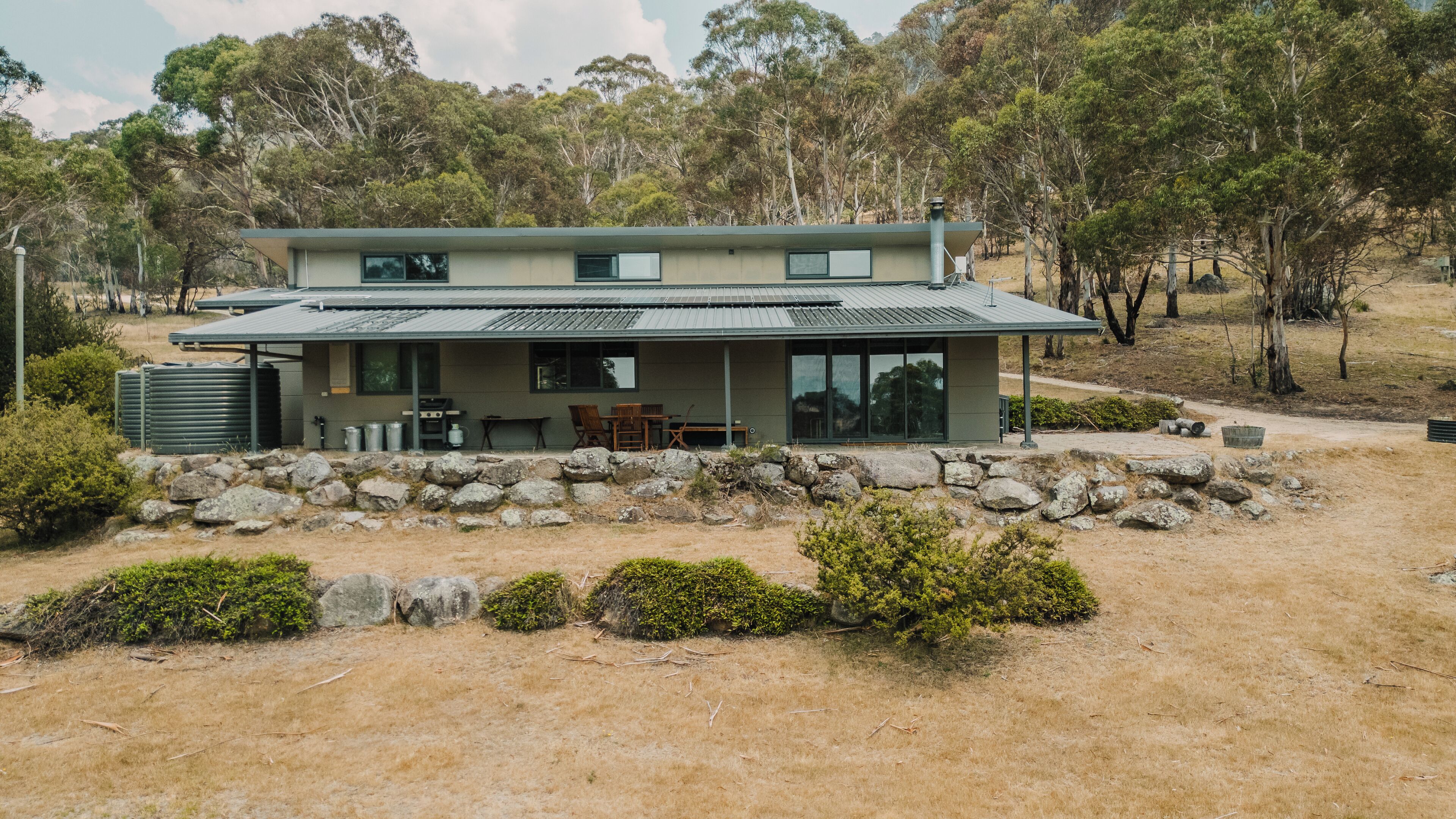 A modern two-story house with a stone wall foundation, large windows, and a covered outdoor seating area.