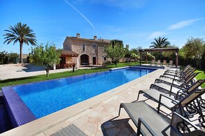 Relax by the serene pool surrounded by a stone deck and lush lawn, with a traditional stone building as a charming backdrop.