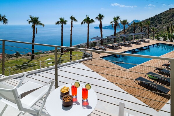 A poolside area with lounge chairs, a table with snacks and drinks, and a view of the sea and hills.