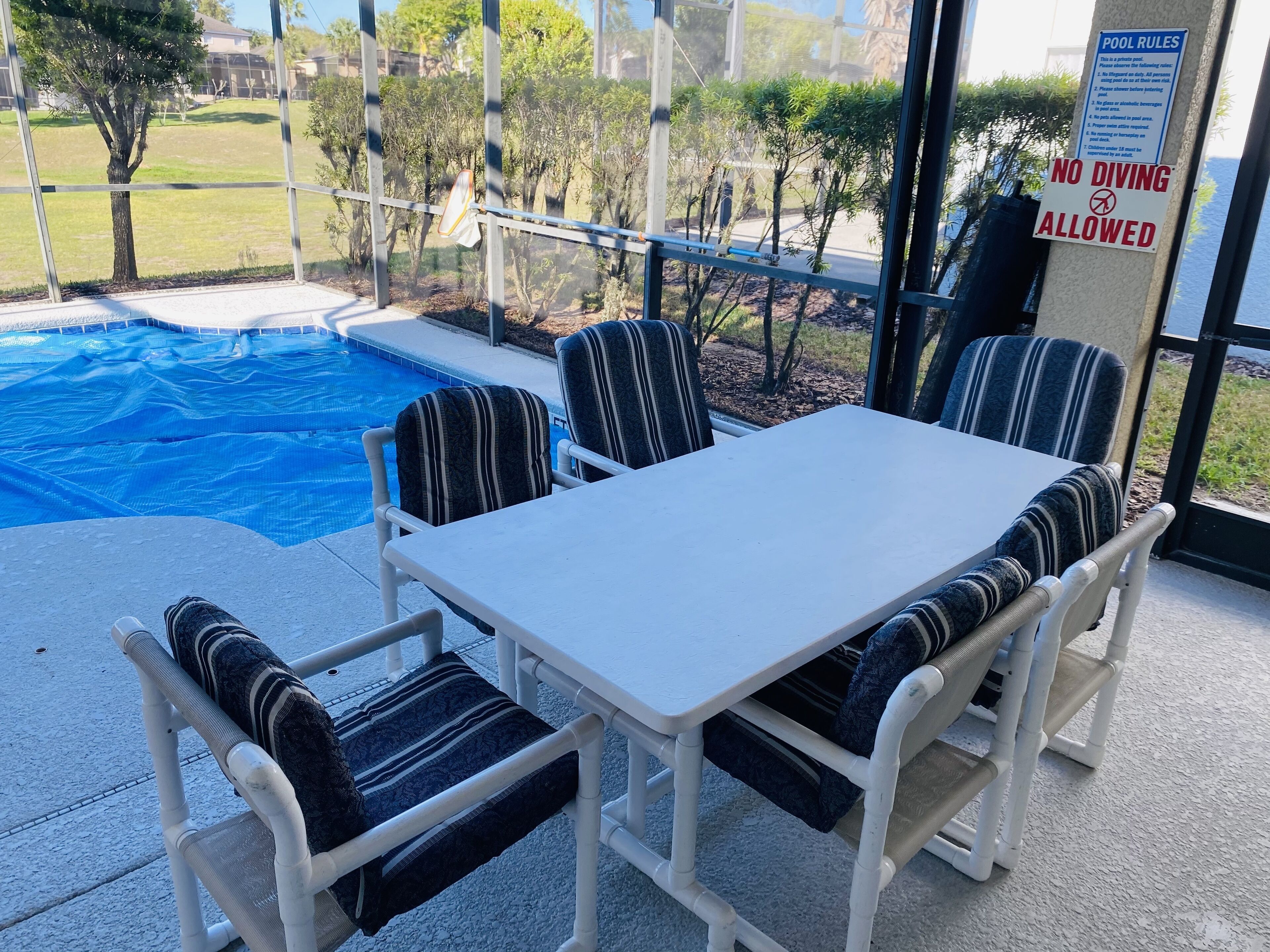 Un espace extérieur avec une piscine, une table et des chaises, un panneau indiquant qu’il est interdit de plonger, et une vue sur le paysage environnant.