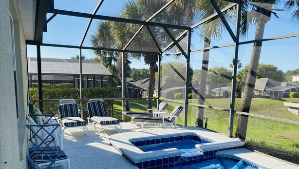 A covered outdoor swimming pool with lounge chairs and a hot tub, surrounded by palm trees and a view of residential buildings.