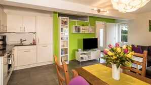 Eine moderne Küche mit weißen Schränken, einem eingebauten Backofen und einer Kaffeemaschine. Es gibt eine Essecke mit einem Holztisch und Stühlen, eine weiße Vase mit Blumen sowie ein Bücherregal mit Büchern und einem Fernseher.