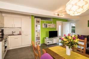 A modern kitchen with white cabinets, a built-in oven, and a coffee maker. There is a dining area with a wooden table and chairs, a white vase with flowers, and a bookshelf with books and a TV.