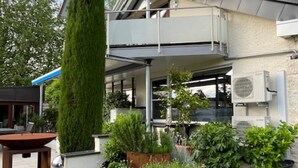 A modern two-story house with a covered patio, a tall coniferous tree, and a circular garden bed with various plants. - Apartment "Zur Imme" with Mountain View, Wi-Fi and Air Conditioning (Lindau)