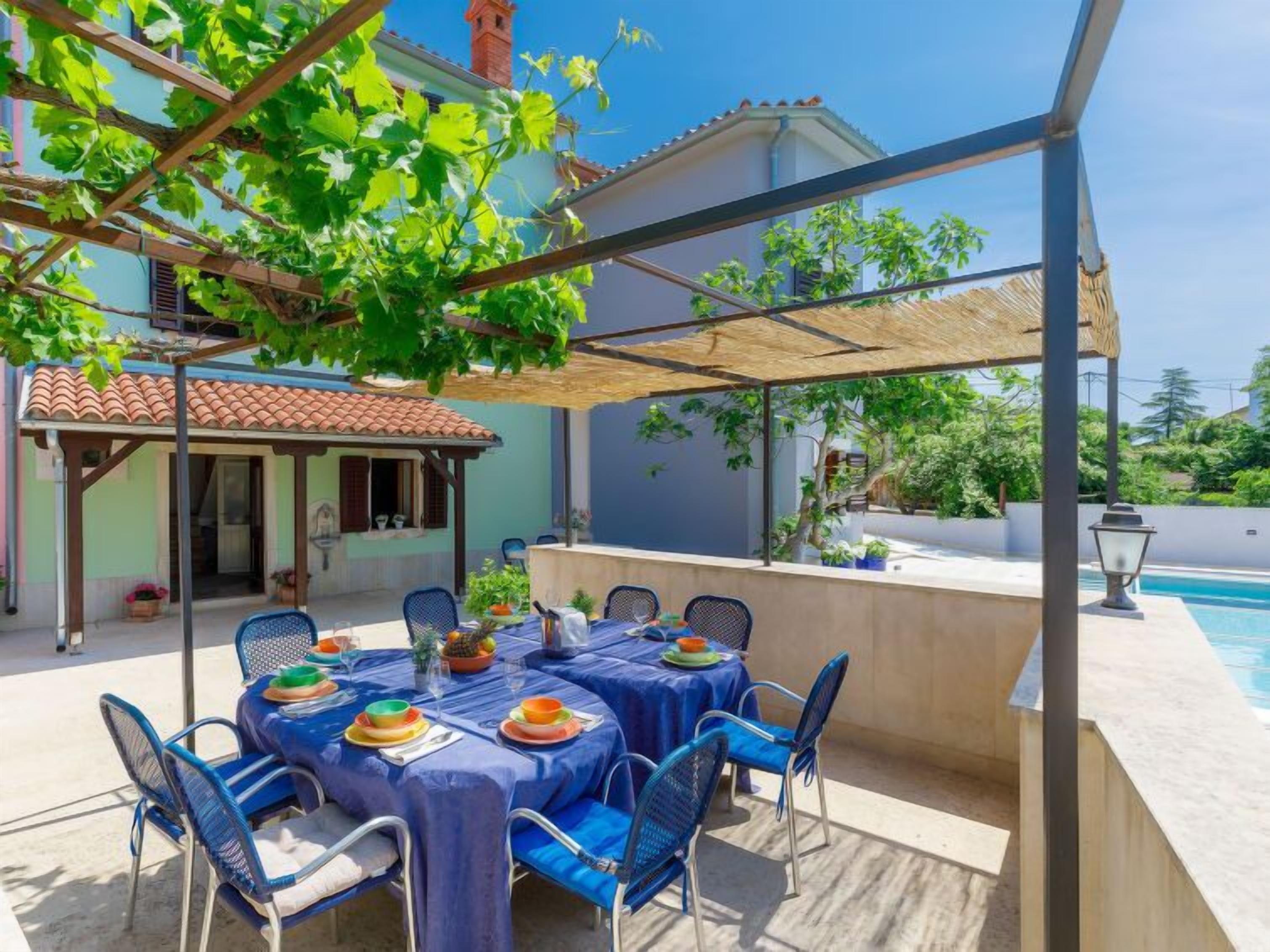 An outdoor dining area with a covered pergola, a pool, and a view of a building with a tiled roof.