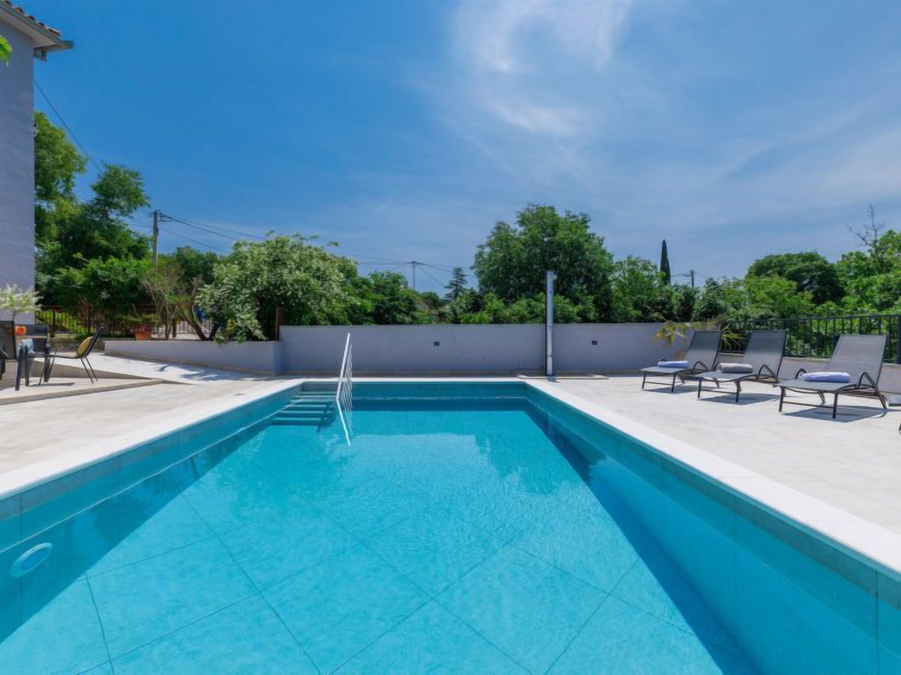 Une piscine rectangulaire avec un escalier permettant d’y accéder, entourée d’une terrasse carrelée de couleur blanche. On y trouve des chaises longues et une petite table.