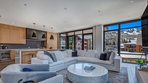 A modern living room with a sectional sofa, a round coffee table, and a view of a snowy landscape through large windows. - Ski-in/out, Best Views, Best Location at Snowmass Base Village | One Snowmass (Snowmass Village)