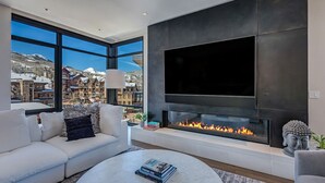 A modern living room with a large TV, fireplace, and a view of a snowy mountain town. - Ski-in/out, Best Views, Best Location at Snowmass Base Village | One Snowmass (Snowmass Village)