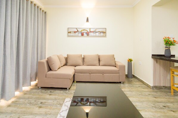 A living room with a beige sectional sofa, a glass-top coffee table, a painting on the wall, and a small plant on a wooden side table. - Marahaba Apartment Hotel (Kampala)