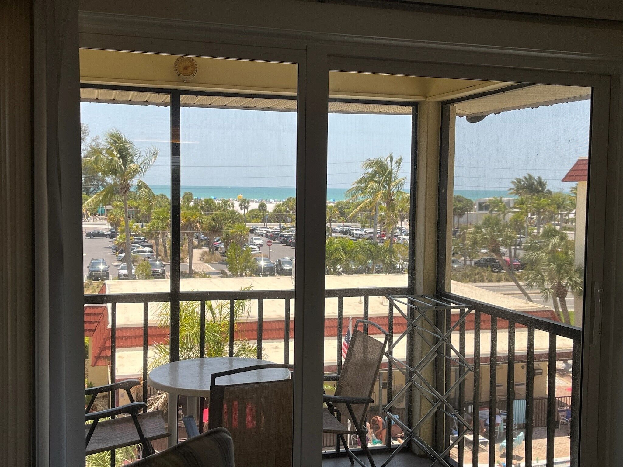 Varanda com mesa e cadeiras, vista para um estacionamento e praia com palmeiras.
