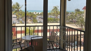 Un balcon avec une table et des chaises, donnant sur un parking et une plage bordée de palmiers.