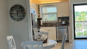 Une cuisine moderne avec des meubles blancs, une table à manger avec des chaises, une horloge au mur et une vue sur la verdure à l’extérieur.
