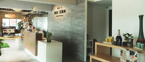 Un espace d’accueil avec une enseigne indiquant « SOJOURN Guest House », un comptoir de réception et un présentoir où sont exposés divers articles.
