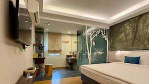 Quarto de hotel moderno com uma cama grande, televisão de tela plana na parede, banheiro pequeno com pia e espelho, e uma janela ampla com vista para árvores.