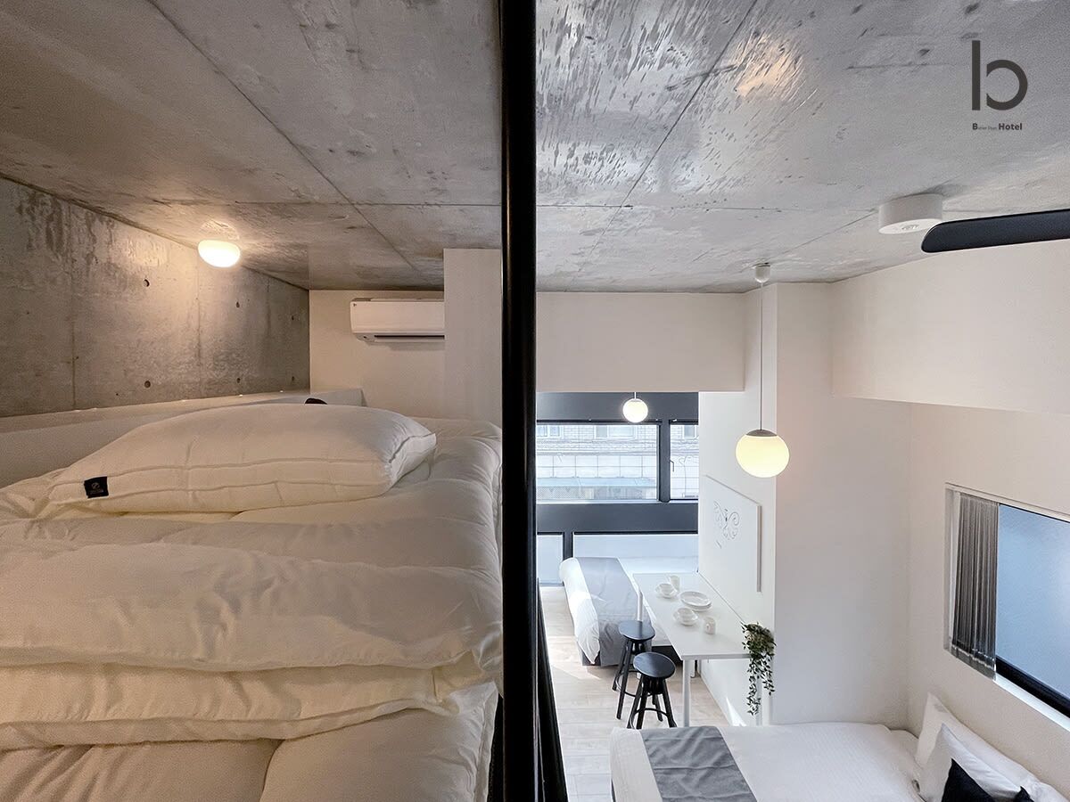 Ett modernt hotellrum med en stor säng, ett litet bord med stolar, en TV och utsikt över staden genom stora fönster.