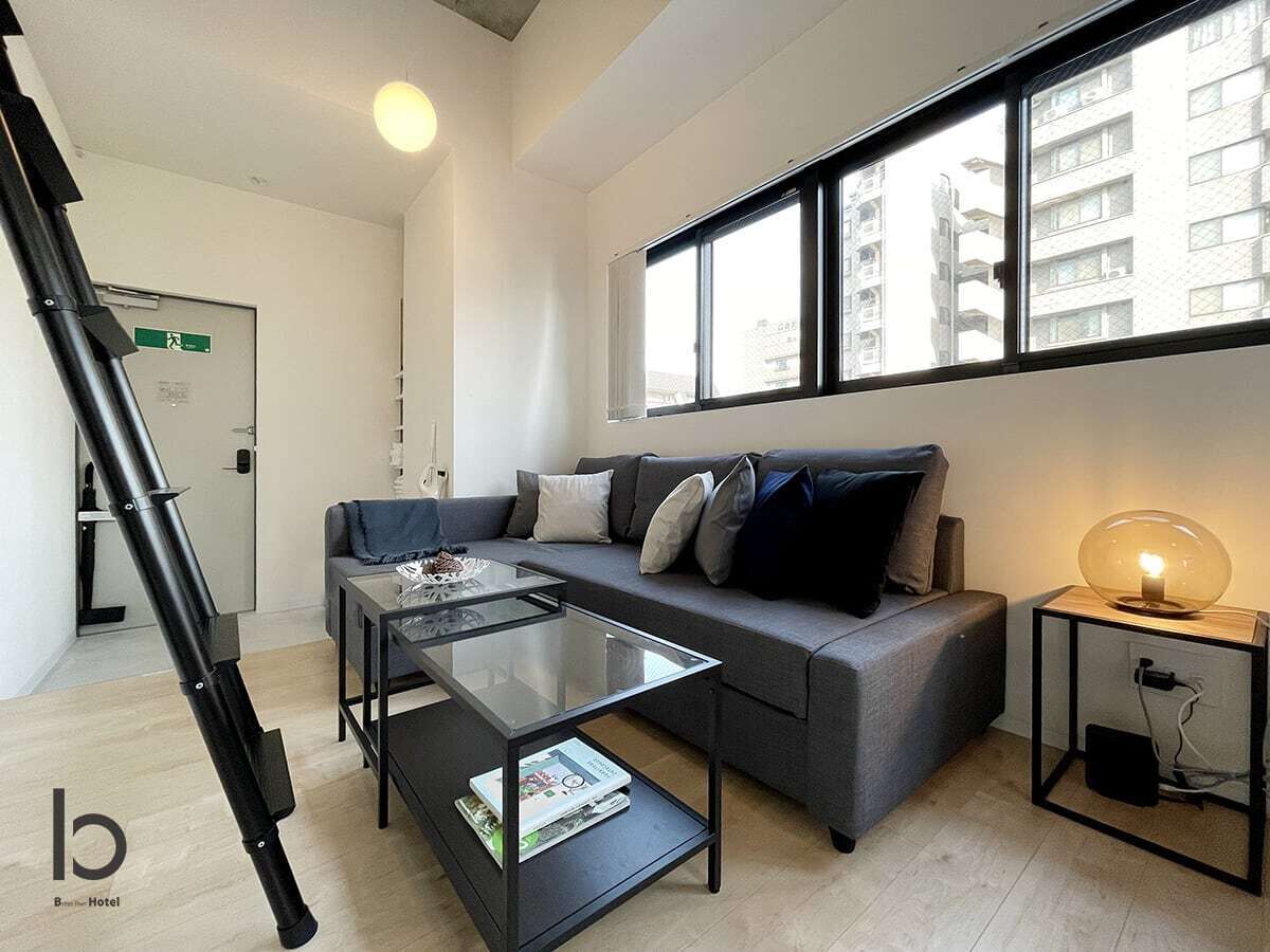 A modern living room with a sofa, coffee table, and a lamp. There is a window with a view of buildings outside.