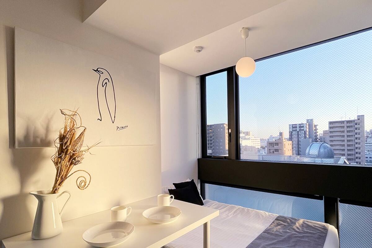 Ein modernes Hotelzimmer mit minimalistischem Design, ausgestattet mit einem Bett, einem kleinen Esstisch mit Stühlen, einem großen Fenster mit Stadtblick und einem an der Wand befestigten Kunstwerk.