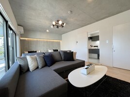 Una sala moderna con un sofá seccional, una mesa redonda de centro y una cocina integrada visible a través de una puerta abierta.