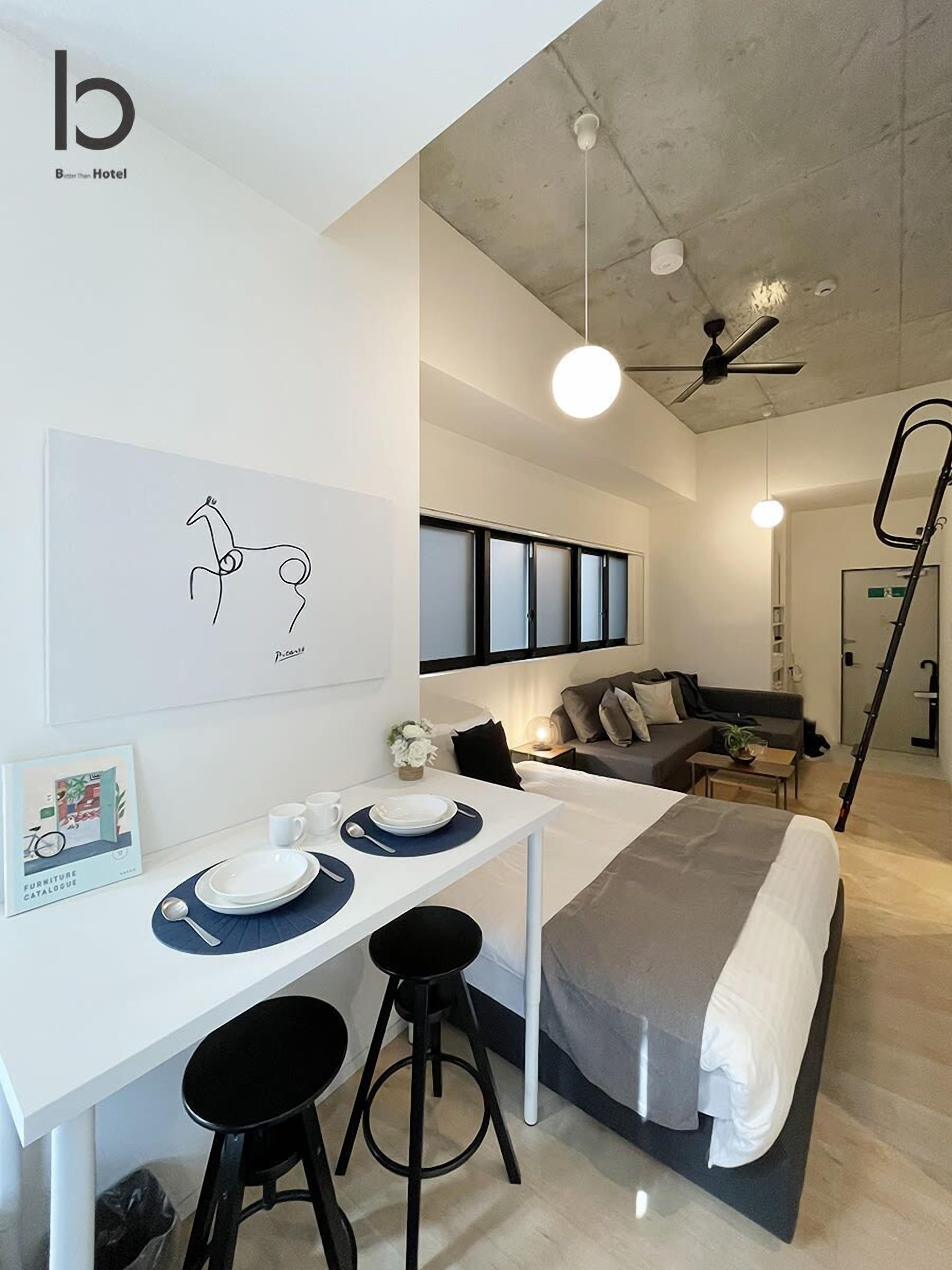 A modern hotel room with a bed, a dining area with a table and stools, a sofa, and a ceiling fan.