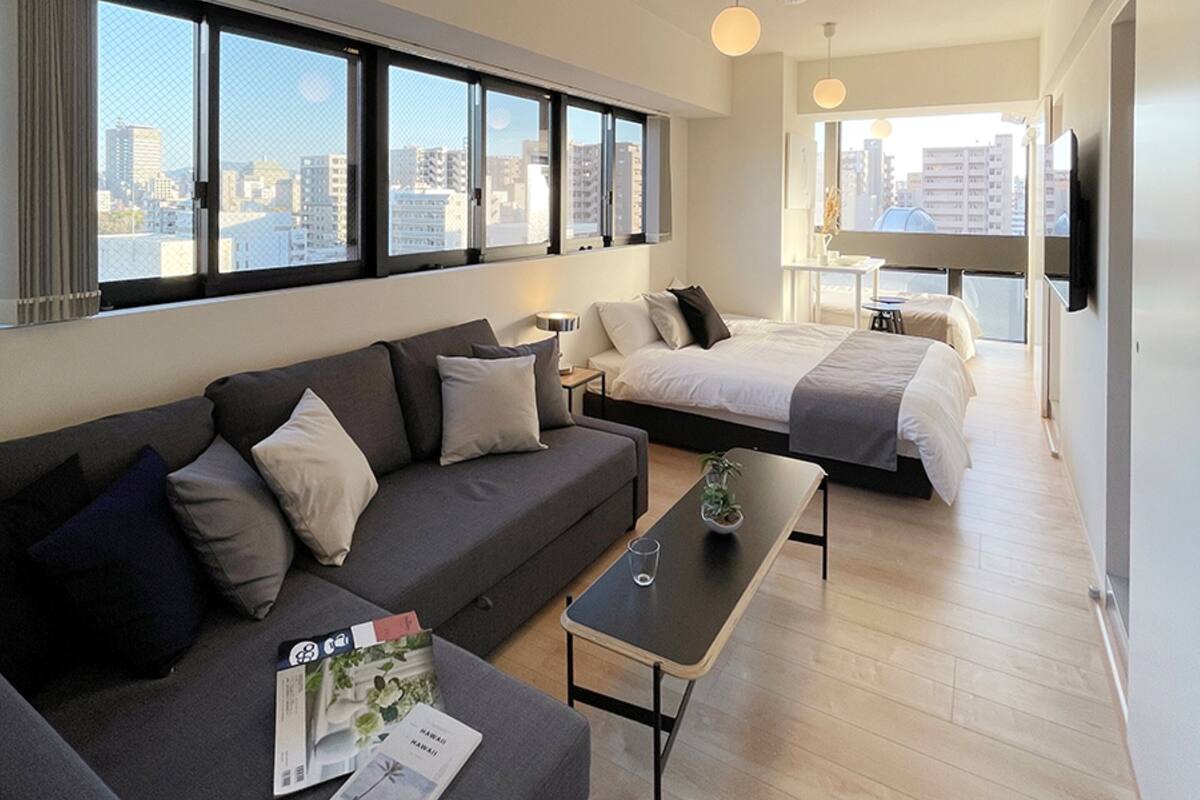 Ein modernes Hotelzimmer mit einem großen Bett, einer Sitzecke und einem Essbereich. Große Fenster bieten Blick auf die Stadt.