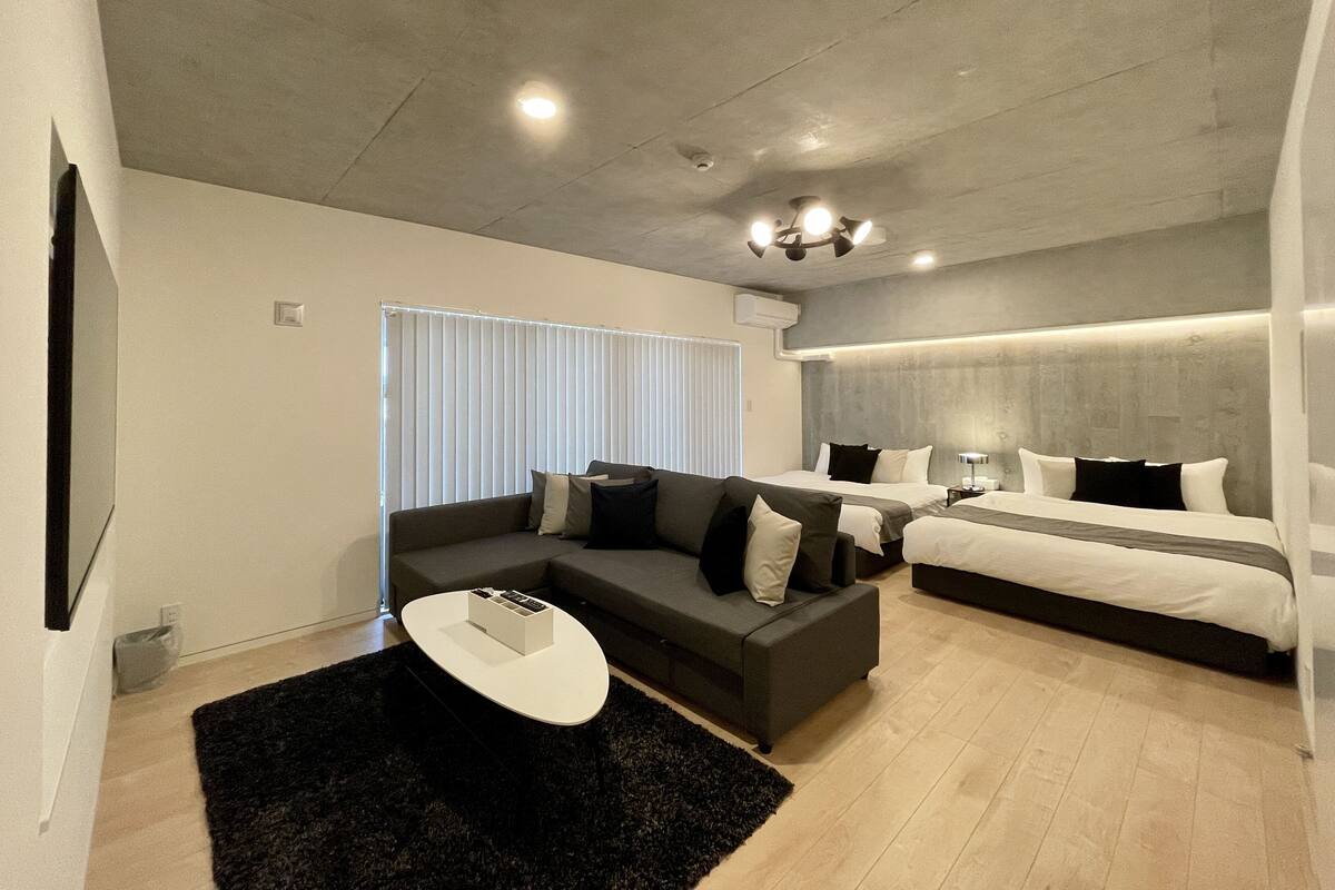 Ein modernes Hotelzimmer mit grauem Sofa, zwei Betten, einem Flachbildfernseher und einer Deckenleuchte.