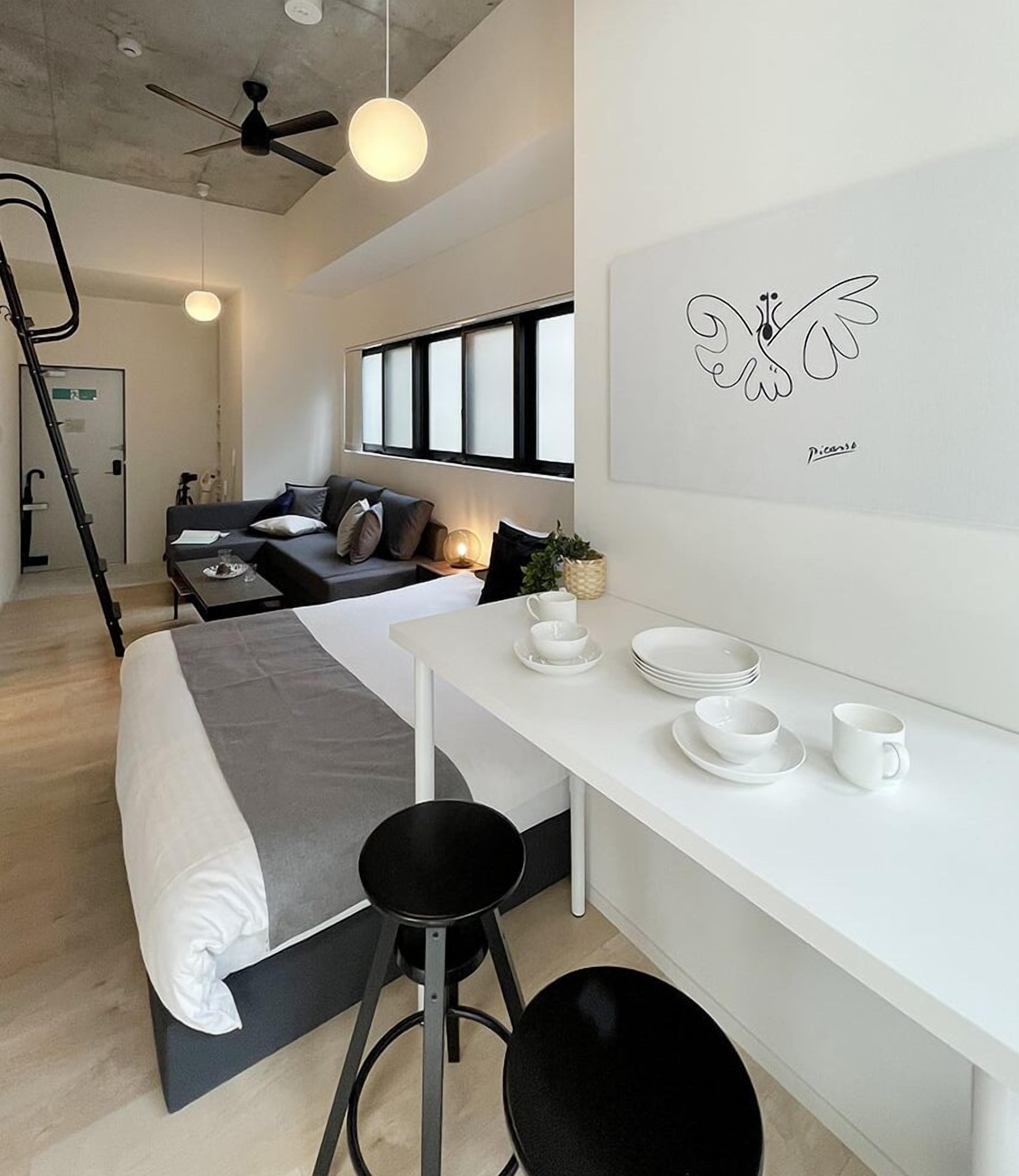 A modern hotel room with a bed, a desk with chairs, a dining table set with plates and cups, a ceiling fan, and a wall with a framed artwork.