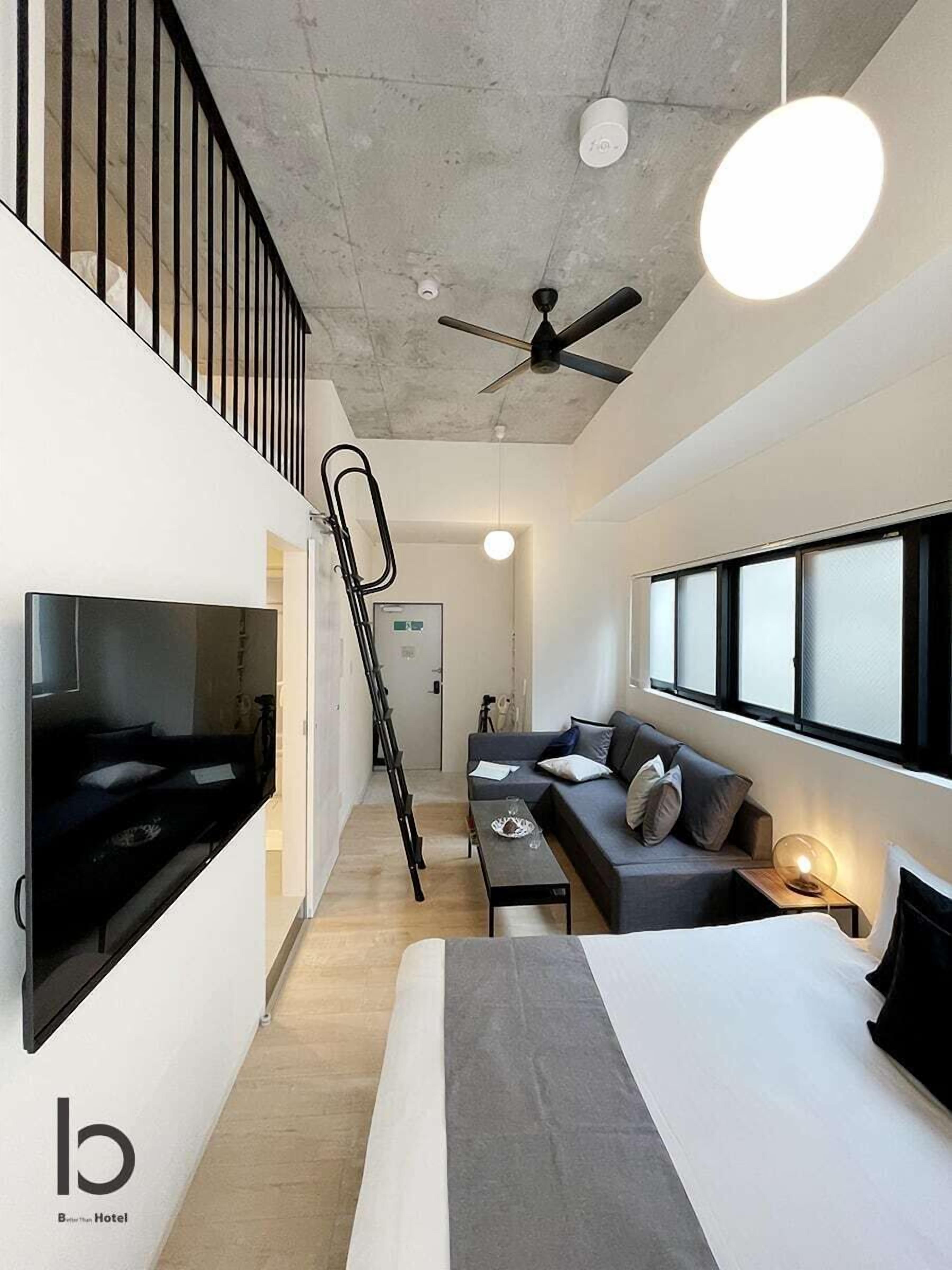 Una habitación de hotel moderna con cama, sofá, televisor montado en la pared, ventilador de techo y ventanas amplias.