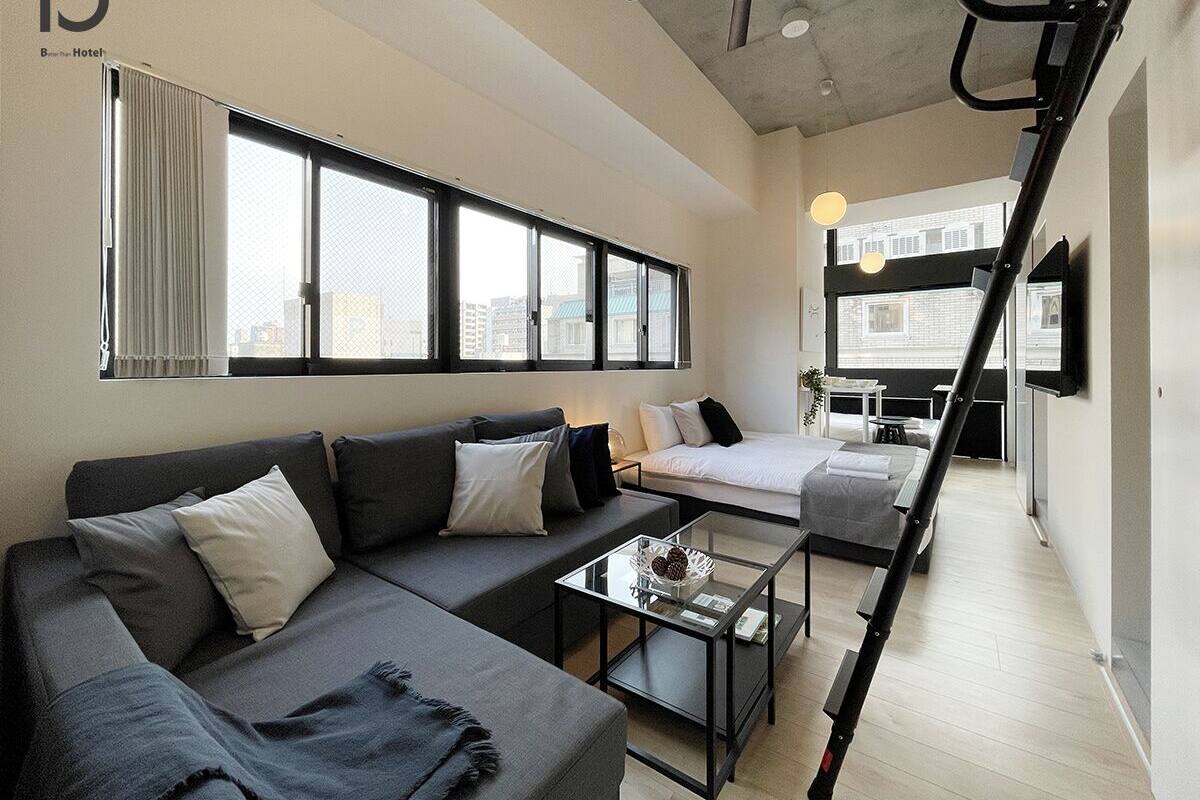 Ein modernes Hotelzimmer mit einer Couch, einem Bett und einem kleinen Tisch. Es gibt große Fenster mit Vorhängen, einen Deckenventilator und einen an der Wand montierten Fernseher.