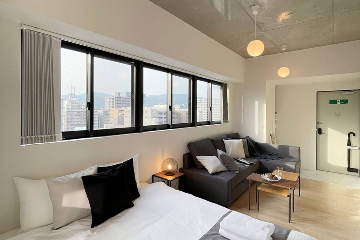 Ein modernes Hotelzimmer mit einem großen Bett, einer Couch, einem kleinen Tisch und Blick auf die Stadt durch große Fenster.