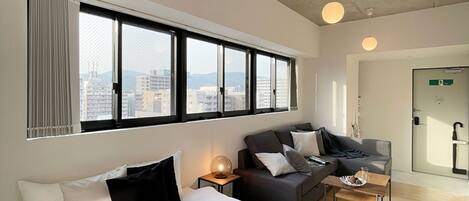 A modern hotel room with a large bed, a sofa, a small table, and a view of the city through large windows.
