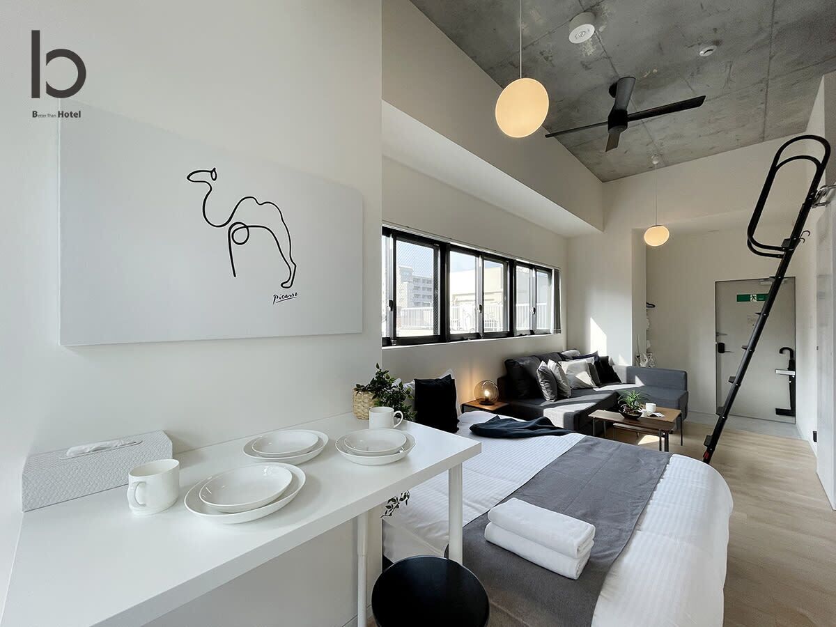 Une chambre d’hôtel moderne au design minimaliste, comprenant un lit, un coin repas avec des assiettes et des tasses, une grande fenêtre et un ventilateur de plafond.