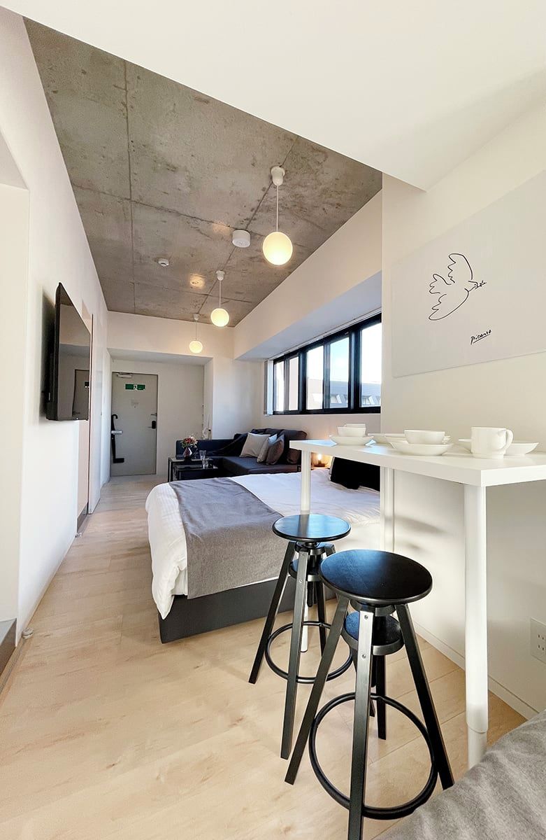 Una habitación de hotel moderna con una cama, una pequeña mesa de comedor, dos taburetes y un ventanal amplio.